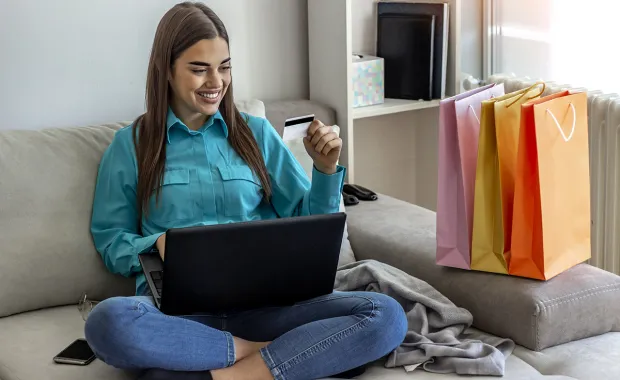 Kvinna sitter i en soffa med shoppingpåsar på soffkanten och betalkort i handen för att shoppa via sin laptop