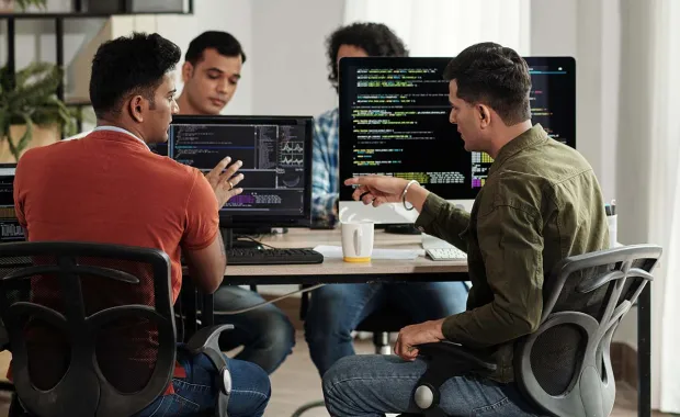 Team members having discussion at desks in front of computers with coding on screen