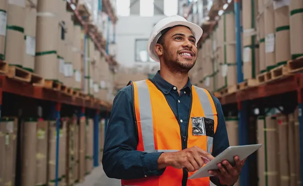 Person wearing a hi-vis vest and hard hat standing in a warehouse using a tablet