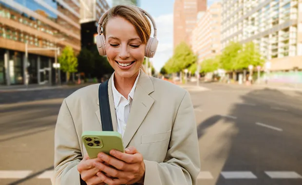 Kuvituskuvassa nuori  onnellinen asiantuntija selaa puhelinta kuulokkeet päässä keskellä kaupunkia