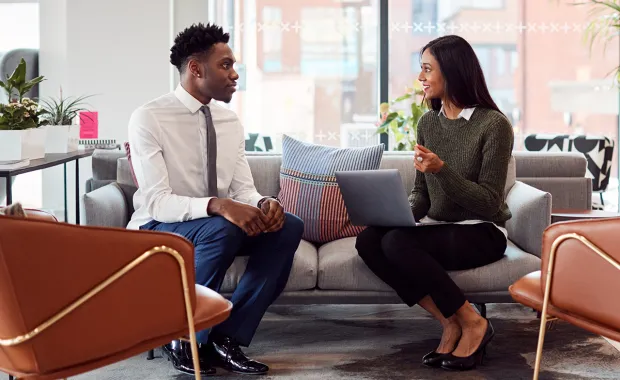 Two people sit on a sofa having a discussion with a laptop