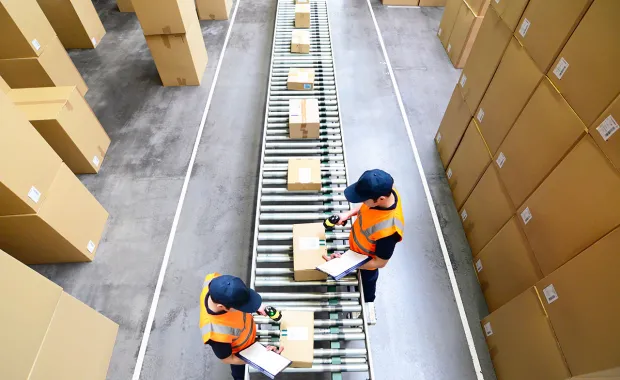 Två lagerarbetare i reflexvästar hanterar paket på ett rullband i ett lager. Stora kartonger är staplade på båda sidor om transportbandet.
