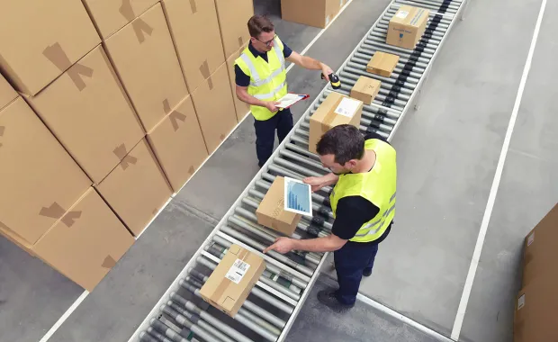 Två lagerarbetare i reflexvästar hanterar paket på ett rullband i ett lager. Kartonger är staplade längs sidorna av transportbandet.