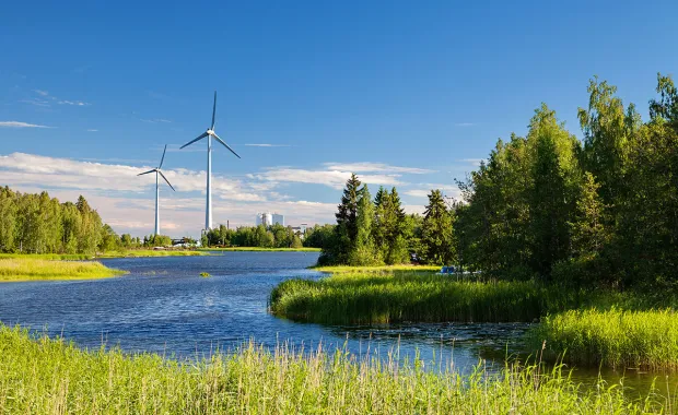 Vindkraftverk står vid en sjö omgiven av grön natur och träd under en klarblå himmel. Bilden visar förnybar energi i ett naturnära landskap.