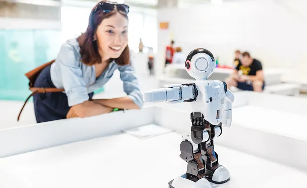 Kvinna står leende framåtlutad mot ett bord i ett öppet kontorslandskap och tittar på en robot