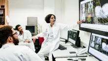Kvinnlig läkare i vit sjukhusrock står lutad mot ett bord och vänder sig mot skärmarna bakom och pekar på patientinformation som hennes kollegor i rummet också tittar på
