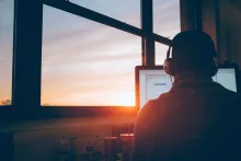 En man ses bakifrån där han sitter på ett mörkt kontor med hörlurar och tittar på en upplyst skärm medan skymningen faller utanför de stora kontorsfönstrena