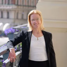 Porträtt av en leende kvinna i kavaj som står vid ett räcke på en balkong eller terrass. I bakgrunden syns stadsmiljö i mjukt fokus, vilket ger ett professionellt och välkomnande intryck.