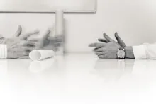 Svartvit bild på två personers händer som sitter mittemot varandra vid ett kontorsbord och ett ihoprullat ark ligger också på bordet