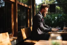 En man med skägg och mörk kostym sitter utomhus i solen vid ett bord och  jobbar vid sin laptop