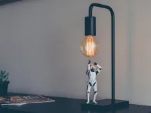 En stormtrooper robot-figur står på en mörk lampfot och tittar upp mot lampan. Lampan står på ett bord och i bakgrunden syns ett magasin