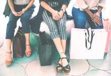 Tre kvinnor sitter på en stor sittpuff i ett shoppingscenter och vilar med shoppingkassor runt sina ben