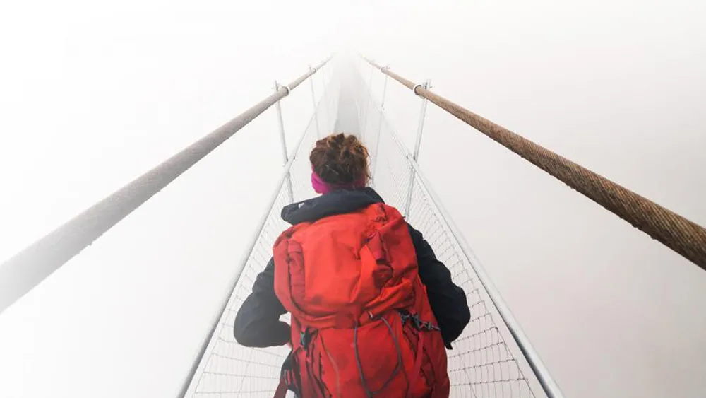 Kvinna med stor röd ryggsäck går över en hängbro i dimma