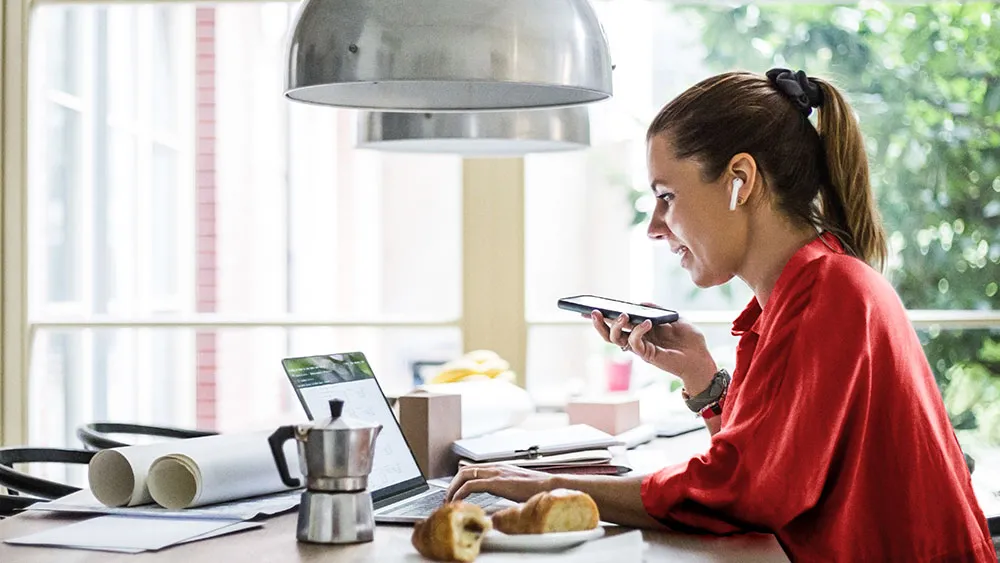 Kvinna i röd topp sitter vid sitt frukostbord i köket och jobbar med laptopen uppfälld och en mobiltelefon i handen