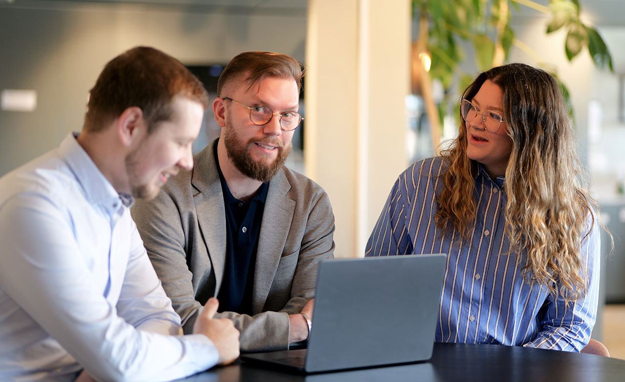 Tre kollegor sitter tillsammans vid ett bord och diskuterar framför en laptop. De ser engagerade ut och samarbetar i en modern kontorsmiljö.