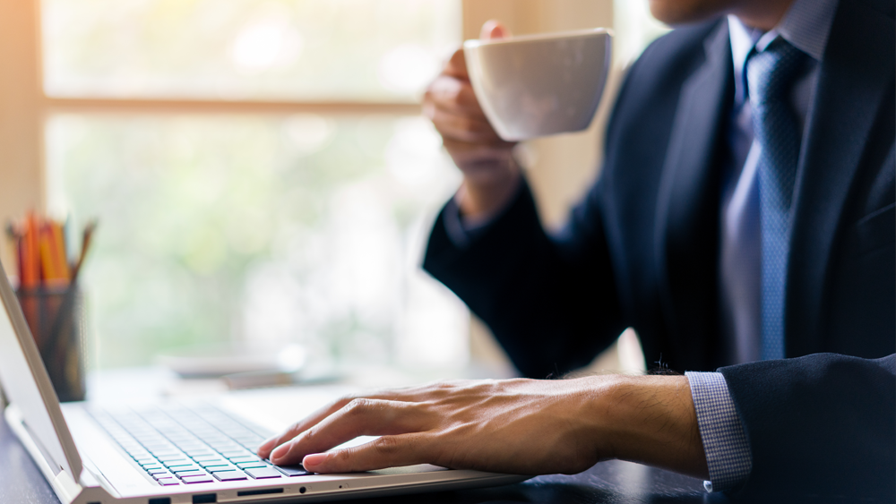 Man i mörk kostym och ljusblå slips sitter med en vit kaffekopp vid sitt skrivbord och läser på sin laptop