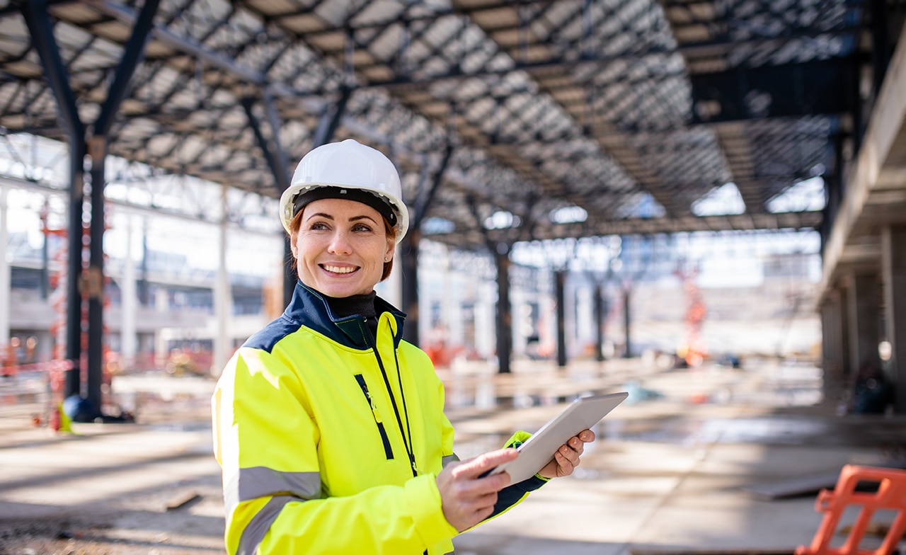 Person med hjelm og refleksjakke holder nettbrett på byggeplass.