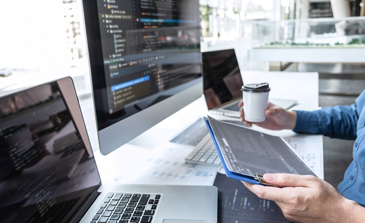 Person holder nettbrett ved arbeidsstasjon med flere skjermer.