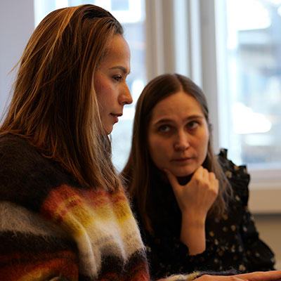 two_women_in_discussion Två kvinnor sitter nära varandra under ett möte. Den ena talar och tittar ner mot en dator, medan den andra lyssnar uppmärksamt med handen under hakan. De befinner sig i ett ljust rum med fönster i bakgrunden.
