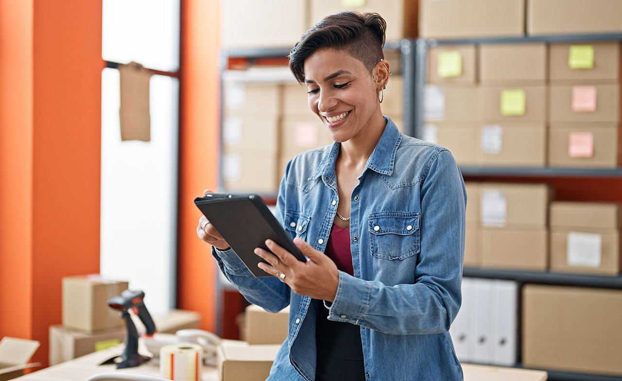 En lagerarbetare står i ett lager och använder en surfplatta för att hantera eller kontrollera inventarier. Hyllor med kartonger syns i bakgrunden.