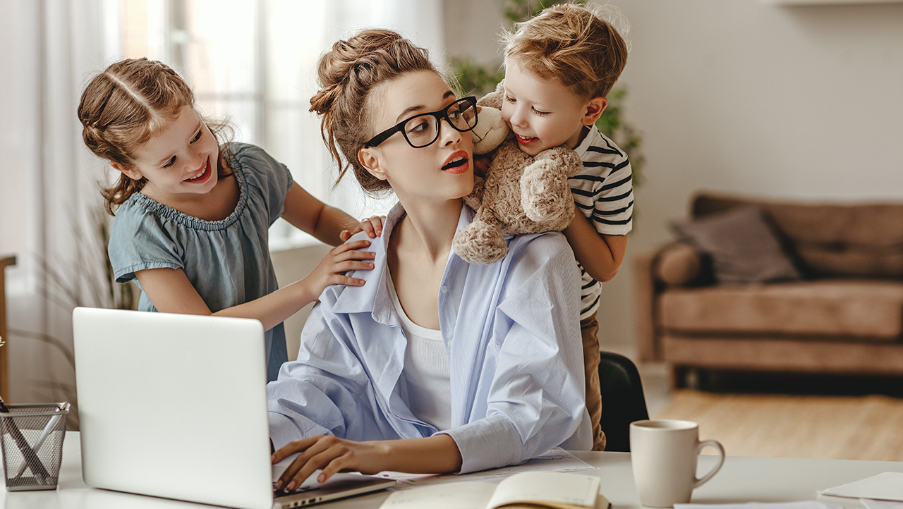 Kvinna sitter framför laptop med två barn bakom sig.