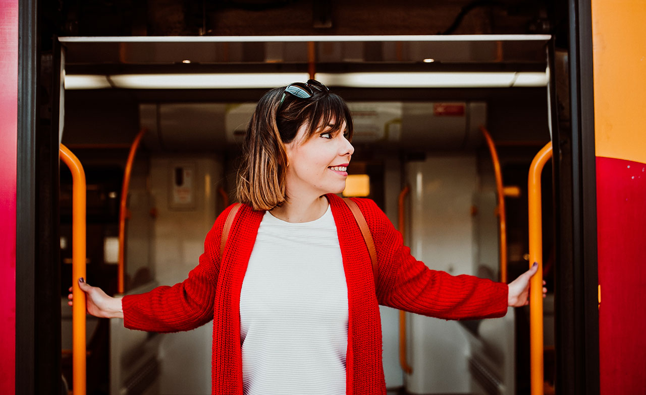 En leende kvinna i röd kappa står i dörröppningen till ett tåg, håller i dörrhandtagen och tittar åt sidan. Hon ser glad och förväntansfull ut, redo att stiga av eller på tåget.