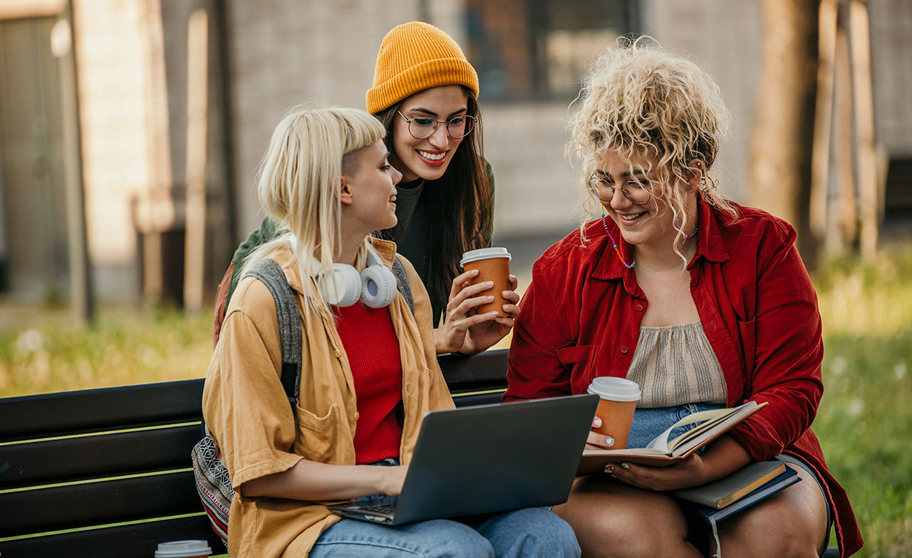 young_women_studying_outdoors_laptop_coffee Tre unga kvinnor sitter på en parkbänk och studerar tillsammans. En har en laptop i knät, en annan håller en anteckningsbok, och alla tre har kaffe i handen. De skrattar och ser engagerade ut i en avslappnad utomhusmiljö.