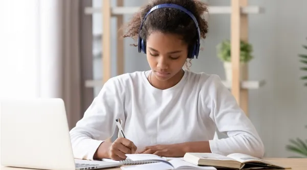 Ung student sitter vid sitt skrivbord hemma och lyssnar på undervisning samtidigt som hon tar…