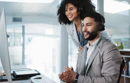 Two office workers wearing headsets look at a computer screen in a modern office
