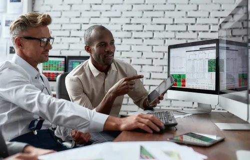 Zwei Experten betrachten gemeinsam Finanzdaten auf Monitoren und einem Tablet.